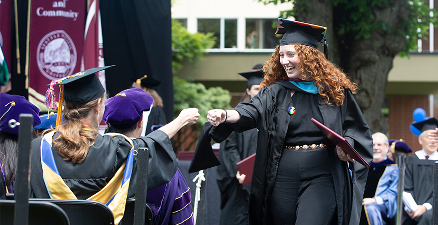 Students in cap and gown fist bump after one receives her graduate diploma at SPU